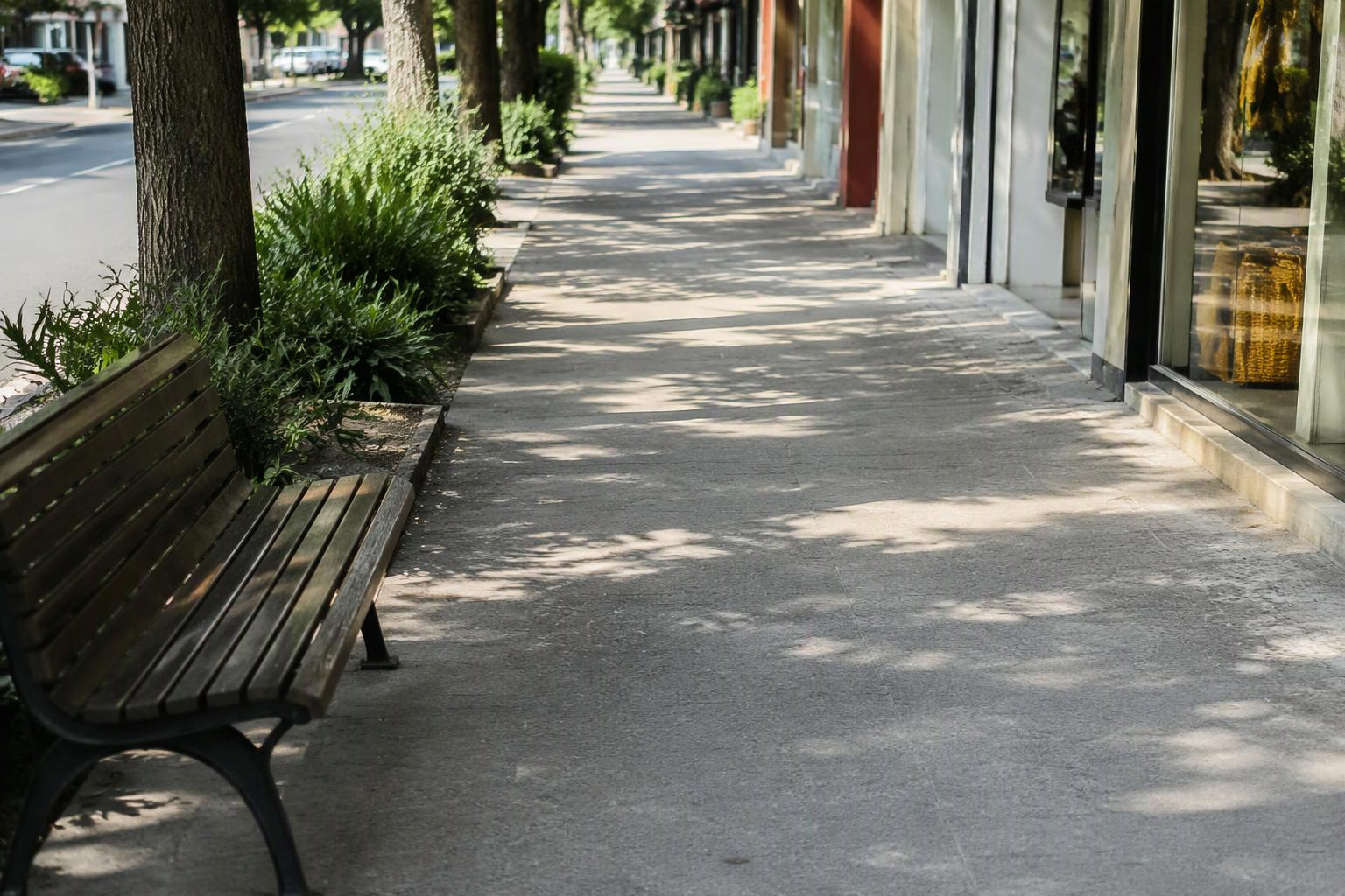 Calçada desobstruída com banco à sombra e estabelecimentos ao fundo.